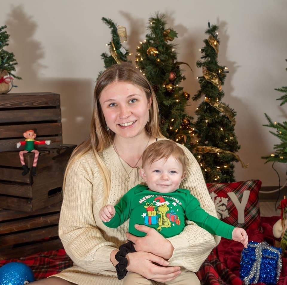 Young mom holding baby boy in green sweater in front of a christmas tree