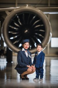Young woman in a flight attendant uniform crouched in front of an airplane beside a toddler in a suit smiling